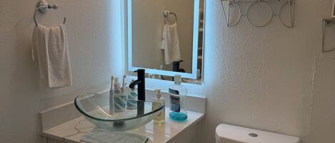 Un baño con un lavabo de vidrio, un inodoro blanco y un espejo con luz superior.