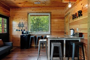 Interior de una cabaña de madera con una zona de cocina, taburetes de bar, una barra y una ventana con vistas a árboles.