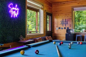 Ein Billardtisch mit Kugeln, eine Neonreklame, eine Mikrowelle und Blick auf Bäume durch die Fenster.