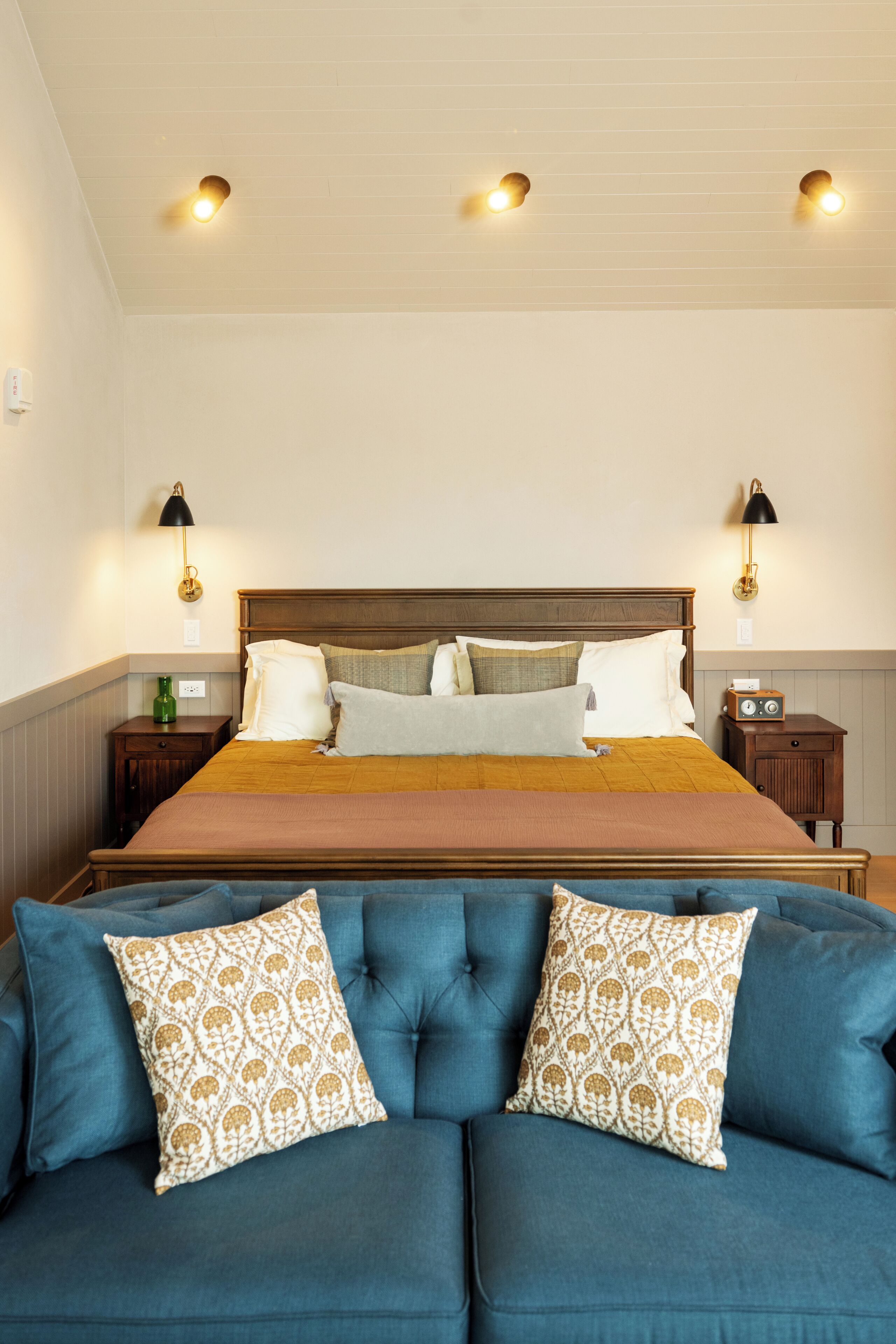 A bedroom with a large bed, two bedside lamps, a nightstand with a radio, and a blue sofa with patterned cushions.