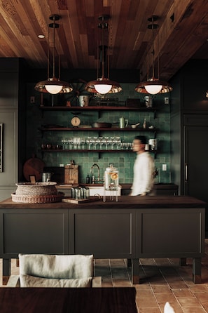 A kitchen with wooden ceiling, green backsplash, and hanging pendant lights.