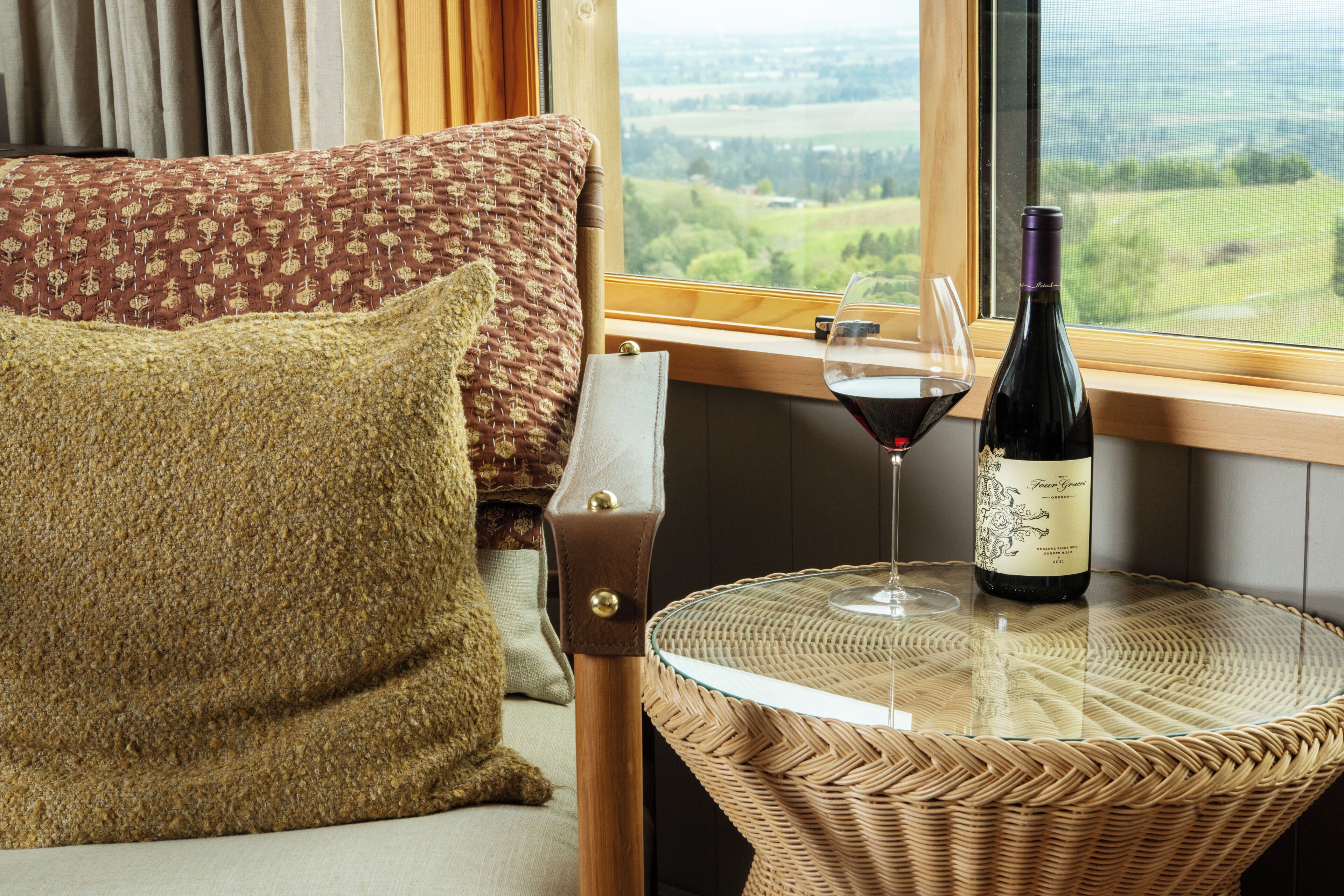 A hotel room with a bed, a pillow, a glass of red wine, and a bottle on a small table by a window.
