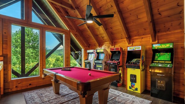 Ein Holzraum mit Billardtisch, Spielautomaten und großen Fenstern mit Blick auf Grünflächen.