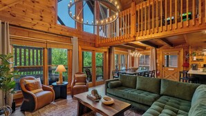 Ein geräumiger Wohnbereich mit einem grünen Sofa, Ledersesseln, einem Holztisch und einem Esstisch mit Stühlen. Ein großes Fenster bietet einen Blick ins Freie.