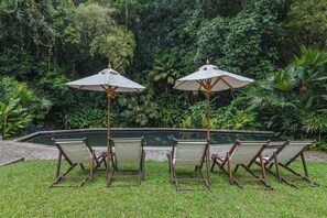 A poolside area with four lounge chairs and two umbrellas, surrounded by lush greenery.