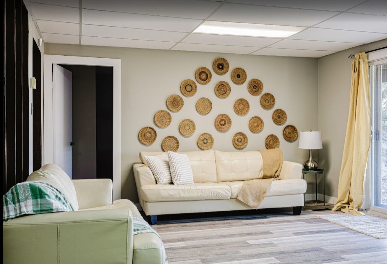 Un salon avec un canapé beige, un fauteuil vert et un mur décoré de motifs circulaires tissés.