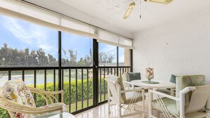 Un balcón con vista a una cancha de tenis y follaje, amoblado con una mesa, sillas y un jarrón de vidrio decorativo.