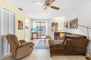 Ein Wohnzimmer mit Sofa, Liege, Deckenventilator, Fernseher, Kunstwerken und Blick durch eine Glastür.
