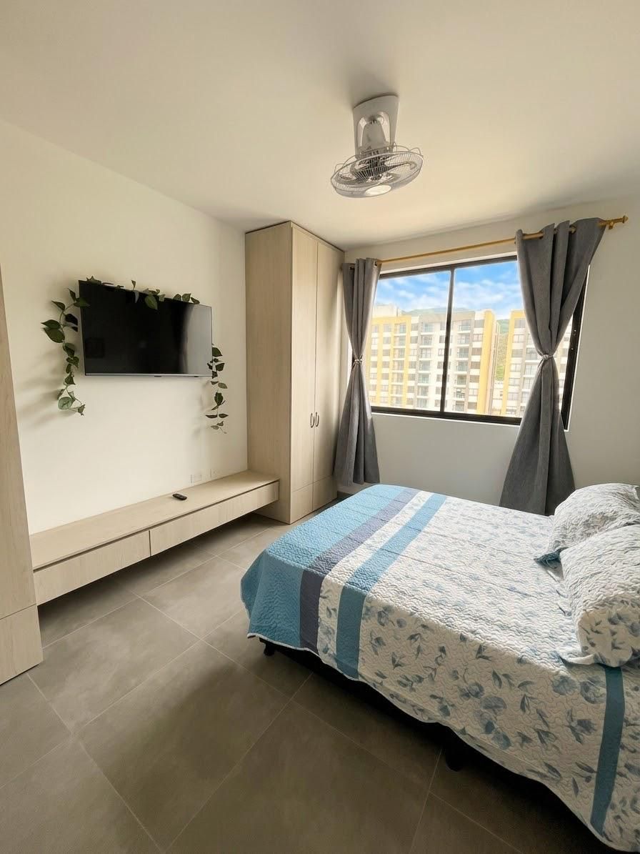 Une chambre à coucher avec un lit, un téléviseur fixé au mur, une fenêtre avec des rideaux et un ventilateur de plafond.