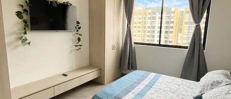 A bedroom with a bed, a TV mounted on the wall, a window with curtains, and a ceiling fan.