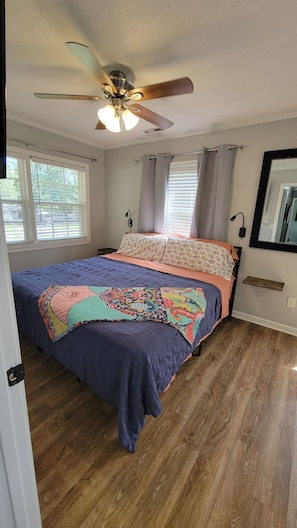 Une chambre à coucher comprenant un lit, un plancher en bois, un ventilateur de plafond, une fenêtre avec des stores et un miroir fixé au mur.