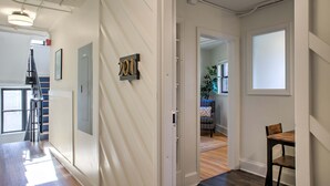 A hallway with a door marked '2011', a staircase, and a view into a room with a plant and a blue chair. - Sabel #201 | Downtown Asheville king suite with parking, Wi-Fi, and smart TV. (Asheville)