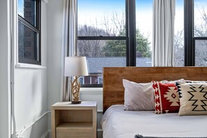 Ein Schlafzimmer mit einem hölzernen Kopfende, einem Nachttisch mit Lampe und Blick durch große Fenster.