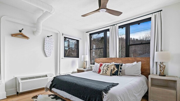 Un dormitorio moderno con cama, mesitas de noche, ventilador de techo y un ventanal grande.