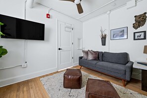 A modern living room with a flat-screen TV, grey sofa, brown ottomans, and a ceiling fan. - Sabel #302 | Downtown Asheville king suite with Wi-Fi, parking, and mini kitchen. (Asheville)