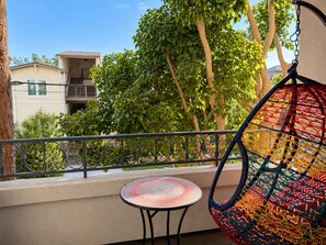 A balcony with a hanging chair, a small round table, and a view of buildings and trees. - Bright 2-bedroom apartment with a large pool by the beach in Marina Del Rey (Los Angeles)