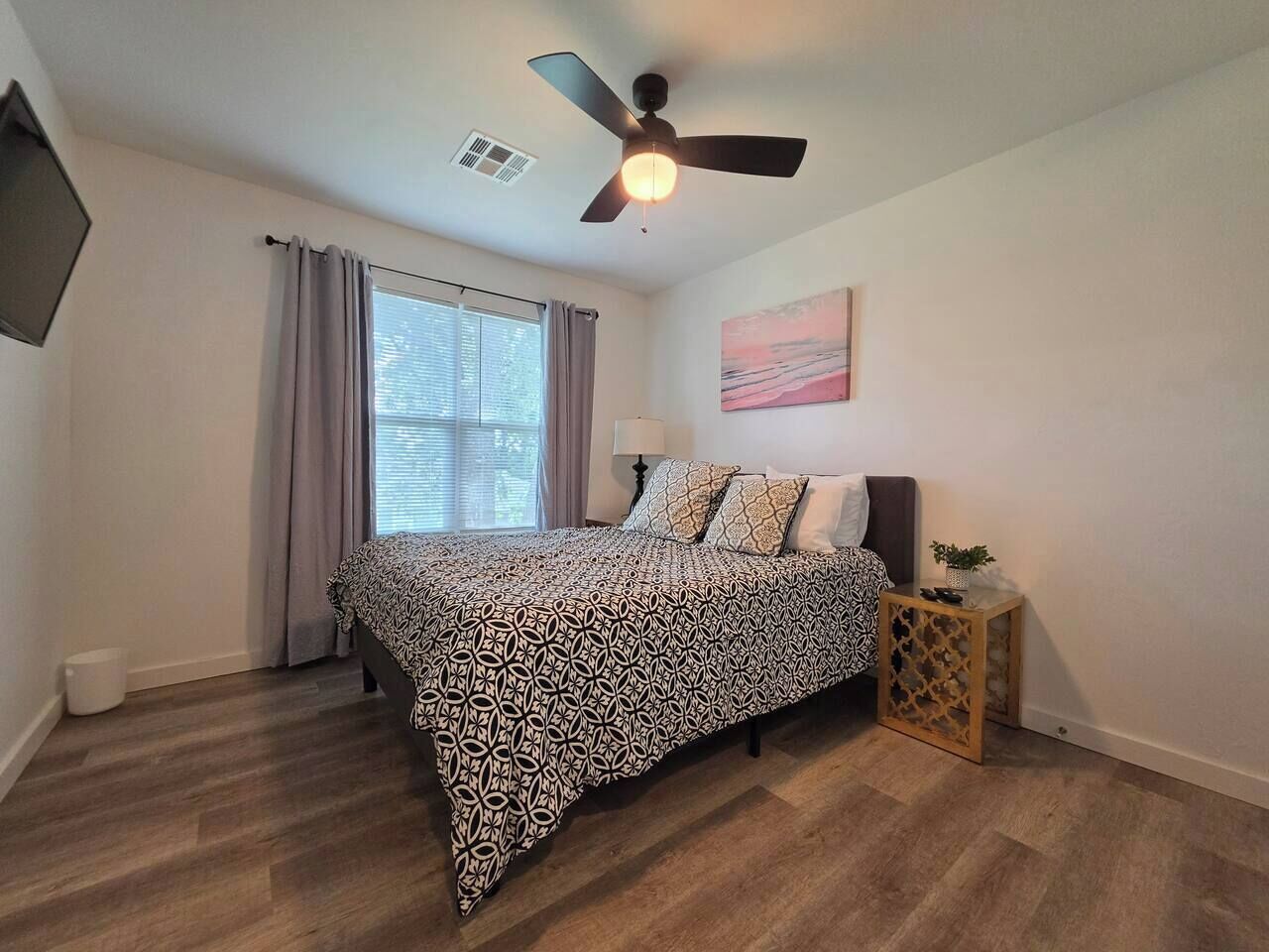 Une chambre à coucher comprenant un lit, un ventilateur de plafond, une fenêtre avec des rideaux, une table de chevet et une œuvre d’art accrochée au mur.