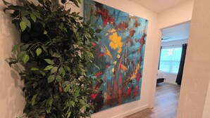 A hallway with a large abstract painting, a potted plant, and a glimpse of a bedroom with a window. - Modern OKC Duplex | Close to OU Med & Downtown (Oklahoma City)