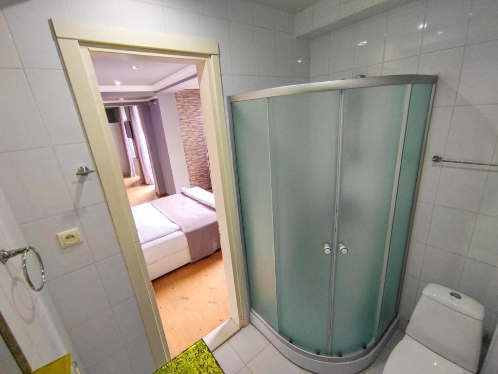 A bathroom with a glass shower enclosure, a toilet, and a visible bedroom with a bed and a brick wall.