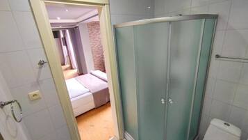 Un baño con cabina de ducha de vidrio, inodoro y habitación visible con cama y pared de ladrillo.