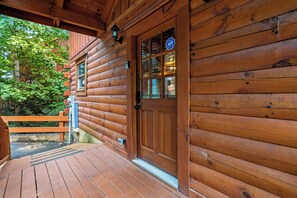 Una cabaña de madera con puerta y ventanas de madera, un porche con barandilla y vistas a árboles.