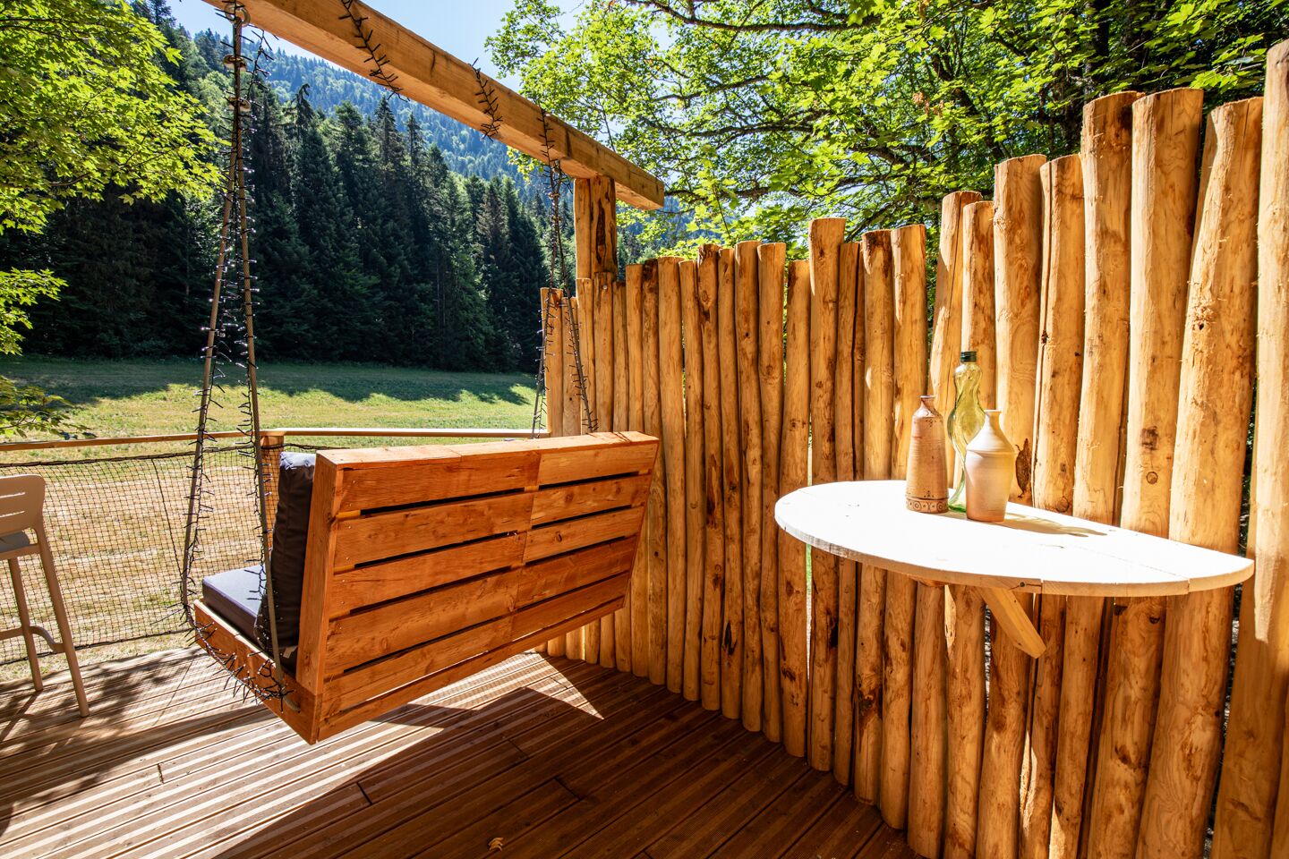 Une terrasse en bois avec une balançoire suspendue, une table ronde avec des bouteilles, et une vue sur une zone boisée.