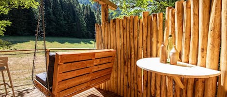 Una terraza de madera con un columpio colgante, una mesa redonda con botellas y vista a un área boscosa.