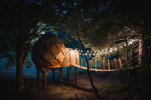 Una casa en el árbol con estructura de madera, rodeada de árboles y un puente colgante.