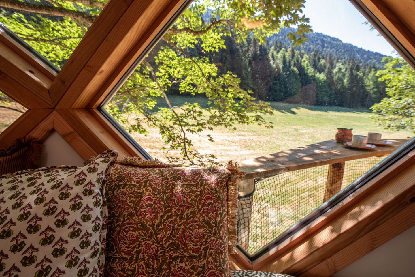 Une pièce dotée d’une grande fenêtre donnant sur une zone boisée, d’un banc en bois et d’une table avec des tasses et une théière.