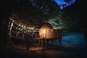 Een houten constructie met een schuin dak, omgeven door bomen en verlicht met lichtslingers.