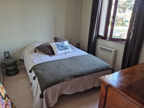 Un dormitorio con cama, almohadas, lámpara de noche, radiador y ventana con cortinas.