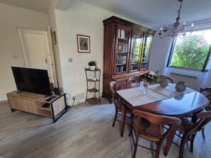 A dining area with a wooden table, chairs, a television on a stand, a bookshelf, and a window with a view of greenery. - Charmante maison à Sundhoffen classée 3 clés parfaite pour des séjours reposants (sundhoffen)