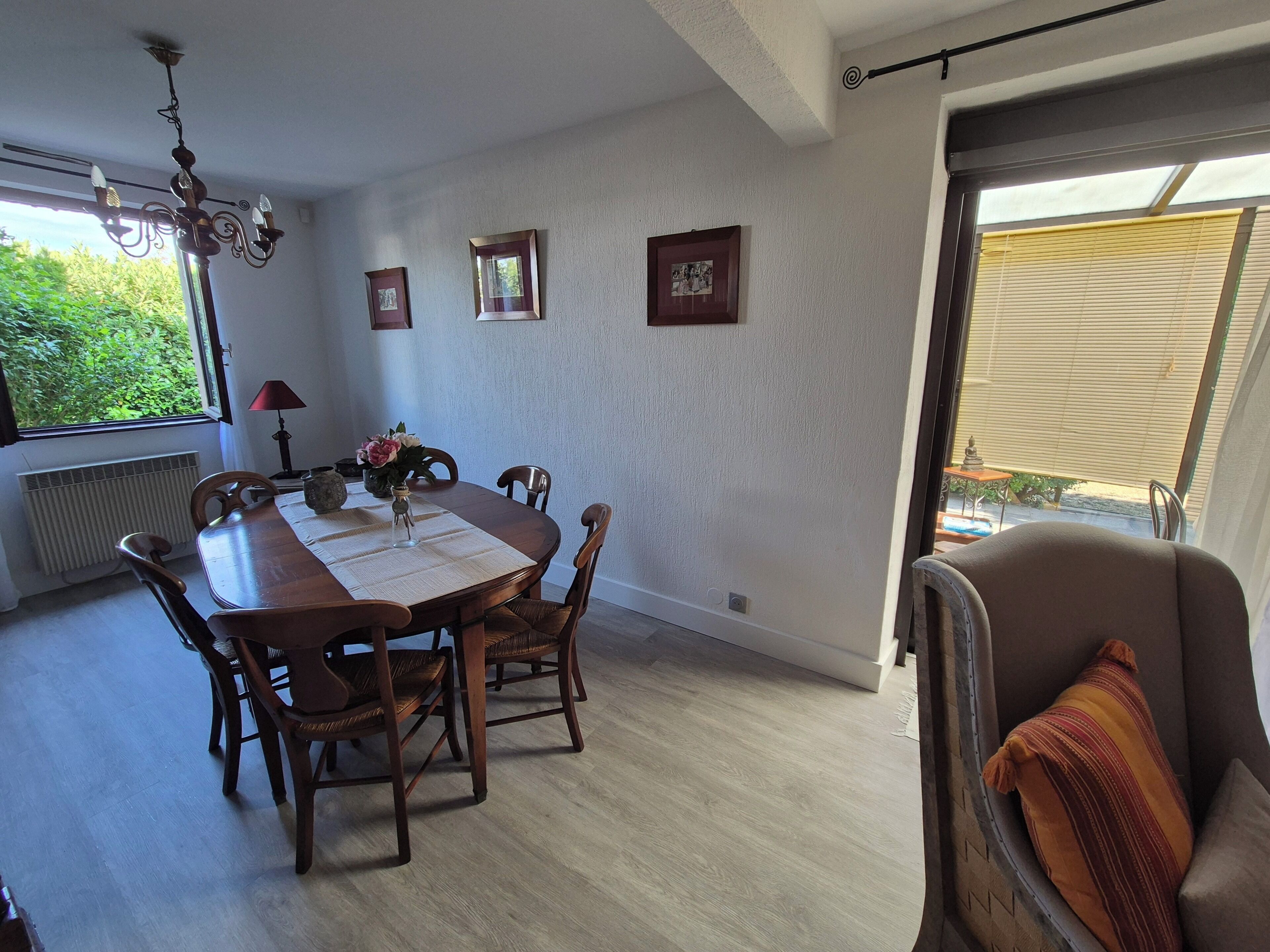 A dining area with a table, chairs, a chandelier, framed pictures, and a view of greenery outside.