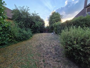 A backyard with a grassy area, dense foliage, and a building in the background. - Charmante maison à Sundhoffen classée 3 clés parfaite pour des séjours reposants (sundhoffen)