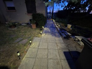 Un sendero pavimentado conduce a la entrada de un edificio, bordeado por macetas con plantas e iluminación exterior.