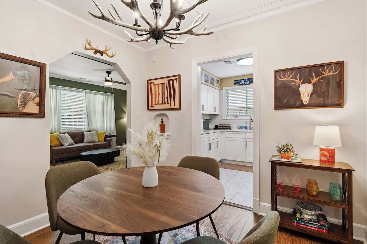 A dining area with a round table, chairs, a vase with dried flowers, a chandelier, and wall-mounted artwork.