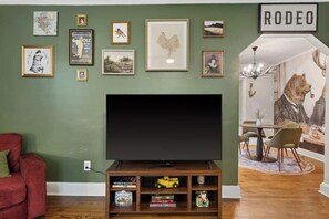 Ein Wohnzimmer mit einem großen Flachbildfernseher, einem roten Sofa, einem Essbereich mit rundem Tisch und Stühlen sowie einer Wand mit gerahmten Bildern und einem 'RODEO'-Schild.