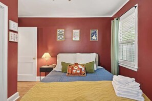 Ein Schlafzimmer mit Bett, Kissen, einem Nachttisch mit Lampe, einem Fenster mit Jalousien und Wanddekorationen.