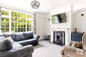 A living room with a grey sofa, a fireplace, a television mounted above it, and a large window with a view of greenery. - 4 Bed House in York, Garden & Private Parking (York)