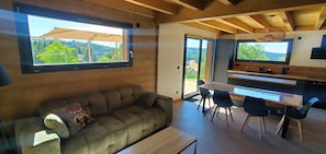 Ein modernes Interieur mit einer Couch, Esstisch, Stühlen und großen Fenstern mit Blick auf Grünflächen.