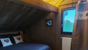 A bedroom with a wooden ceiling, a bed with a blue bedspread, two heart-patterned pillows, a window with a view of greenery, and a curtain.