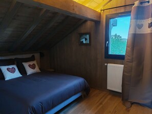 A bedroom with a wooden ceiling, a bed with a blue bedspread, two heart-patterned pillows, a window with a view of greenery, and a curtain. - Sublime chalet au coeur des montagnes vosgiennes - 3 chambres, Sauna, Jacuzzi (Anould)
