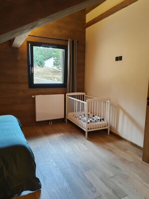 Ein Zimmer mit Holzboden, einem Reisebett, einer Heizung und einem Fenster mit Blick auf Grünflächen.