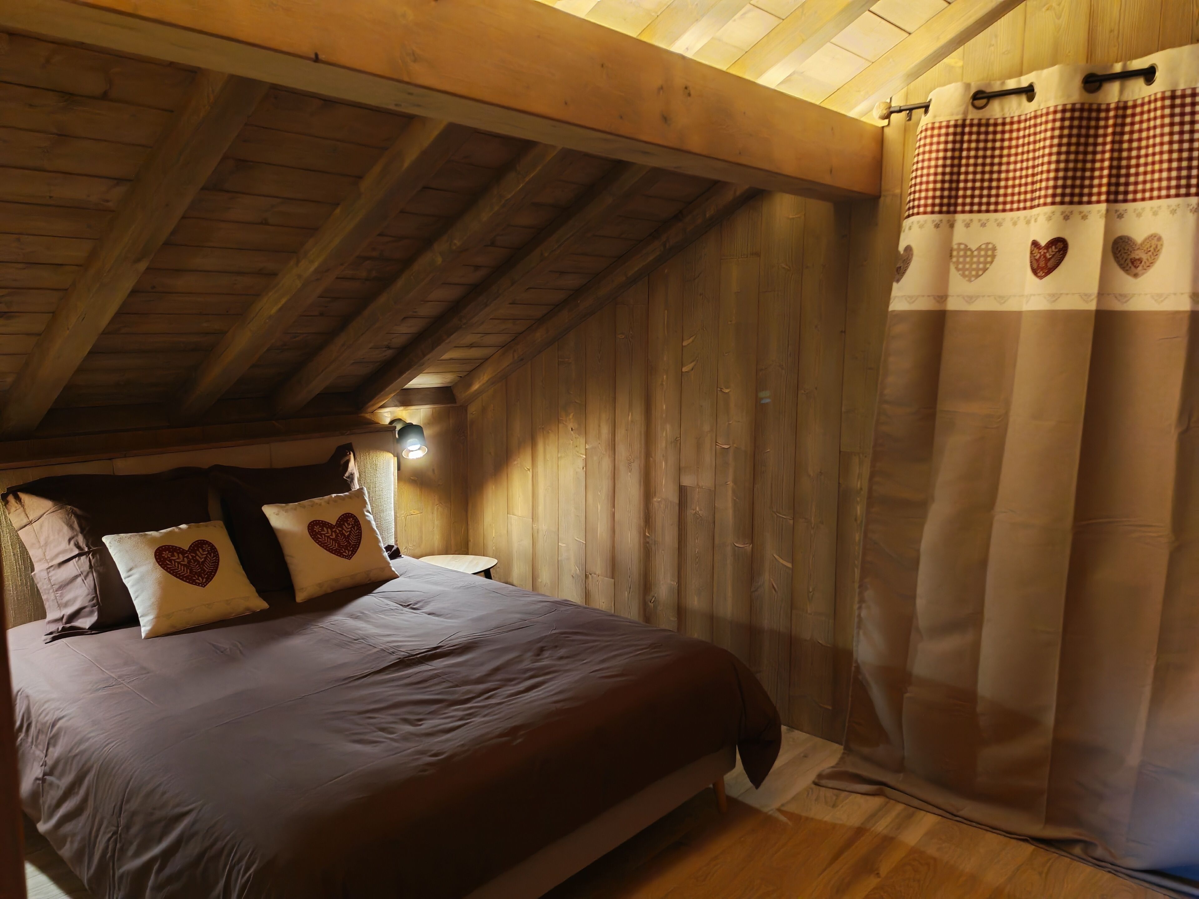 Un dormitorio con techo de madera, cama con almohadas estampadas con corazones y cortina a cuadros.