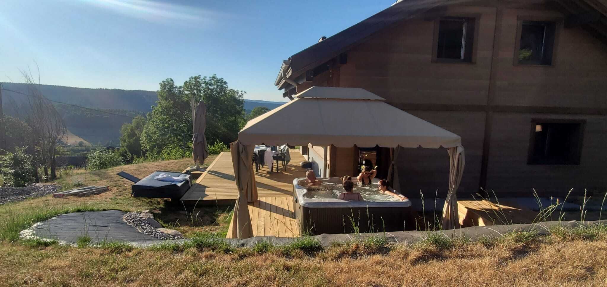 Et område med jacuzzi, træterrasse og et telt, omgivet af et naturligt landskab.