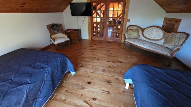 Um quarto de sótão acolhedor com soalho de madeira, uma televisão na parede, um sofá, uma cadeira e duas camas.