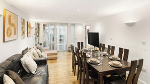 A dining area with a wooden table set for four, a leather sofa, and a TV on the wall. - Exquisite 4BR Home 15 Guests Prime Central London (London)