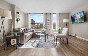Ein modernes Wohnzimmer mit Sofa, Couchtisch und Sesseln, einem großen Fenster mit Blick auf die Stadt und einem Flachbildfernseher an der Wand.