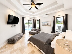 A modern bedroom with a bed, a flat-screen TV, a ceiling fan, and a view of the outdoors. - Spacious 5-bedroom Luxury Courtyard Estate in ALVA Pool + Resort Style Amenities (Alva)