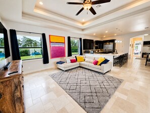 A modern living room with a sectional sofa, a kitchen in the background, and a large window with curtains.